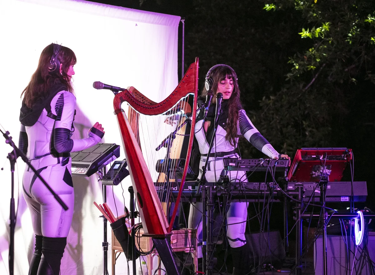 Sarah Glass and Alisa Rose Healy in white space suits at their drum pads, synths and a red harp under violet stage light