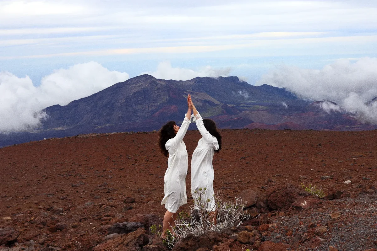 Sarah Glass and Alisa Rose Healy in white, hands joined above their heads, on the red cinder summit of Haleakalā with clouds and the crater rim behind them
